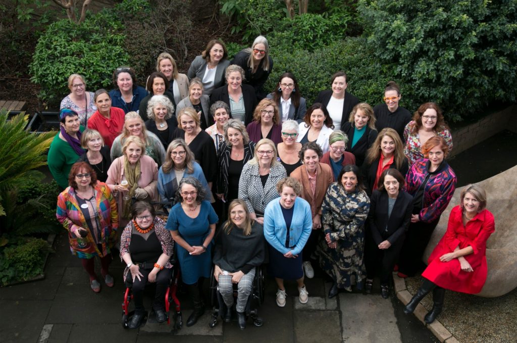 A group shot of the many women who came along to the 2023 Take on Board Alumni Dinner. The group are huddled together in the lovely University House gardens, looking up as photographer Shaney takes the picture from the second floor window.