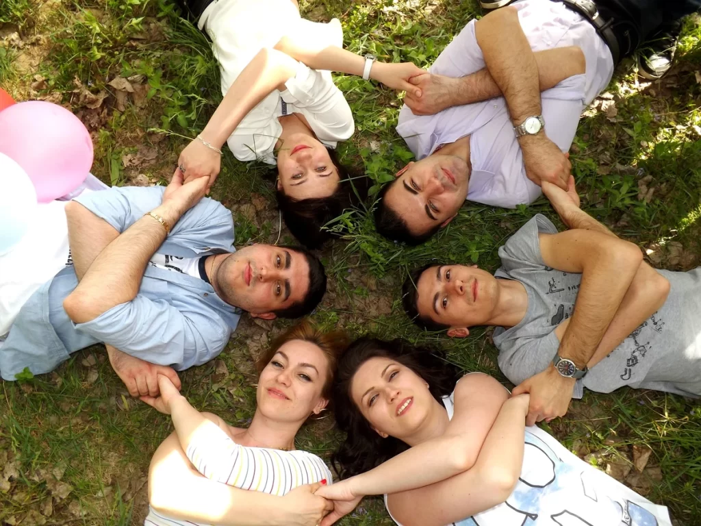 Six people are lying on the grass, facing upwards, heads pointing inside the circle. Their shoulders are touching and their arms are interlaced and held together by their hands. They are smiling.