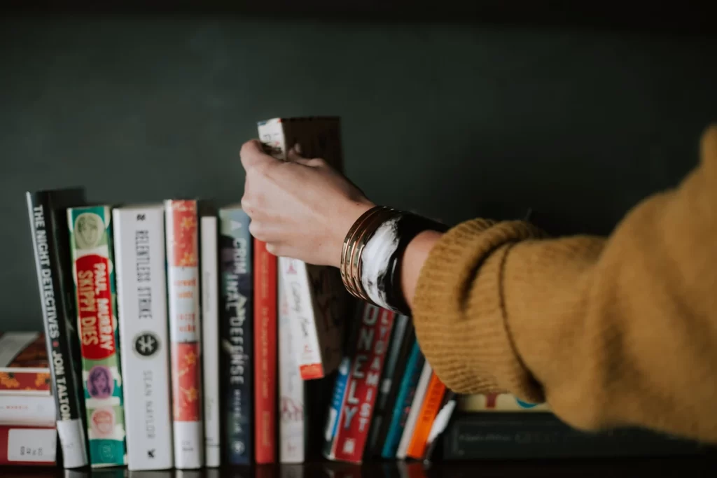 A woman's hand reaches into a bookshelf with around 20 books.. Her hand has hold of one book, lifting it into the air. We can on see her arm - a mustard jumper and a few bangles.