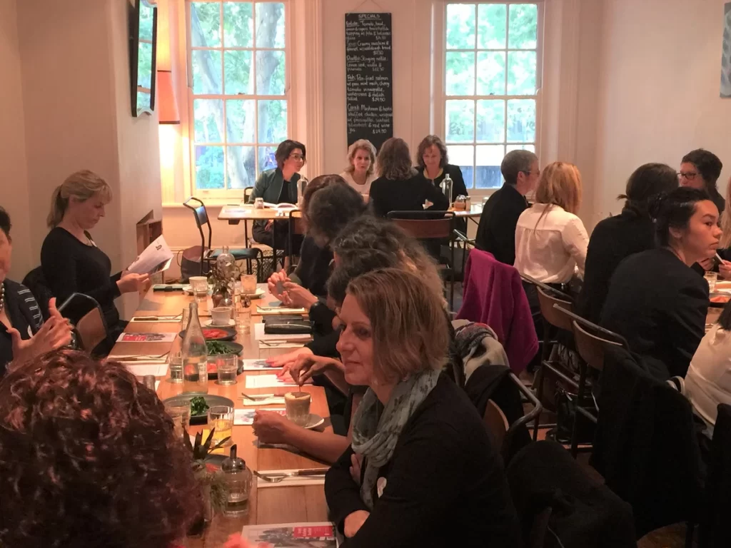 Two long tables of women attend the December 2017 Take on Board breakfast. The three presenters sit at a table at the front of the room, under windows.