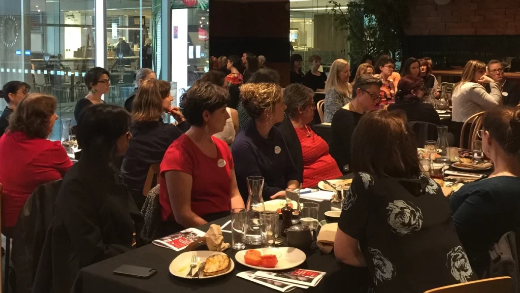 A room full of women seated at long breakfast tables all look to the front of the room at the May 2018 breakfast, listening intently to the presenters.