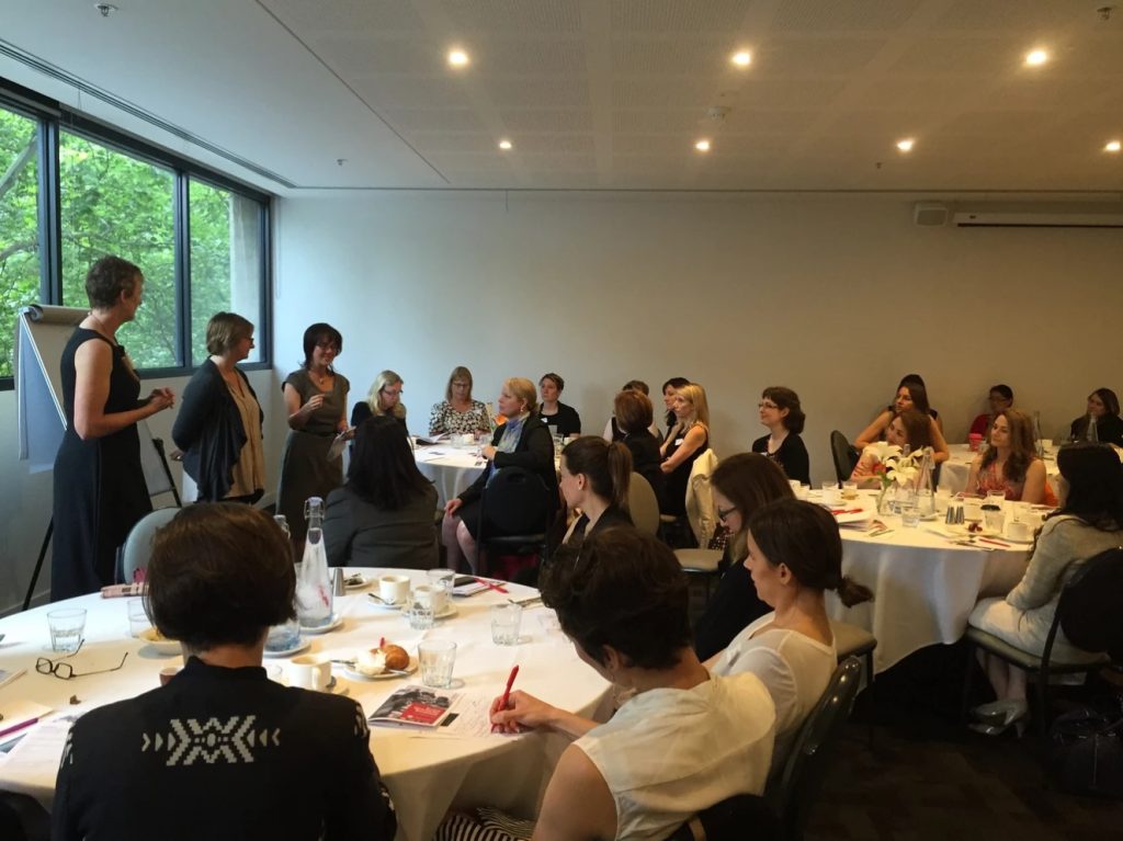 Women sit around three round tables listening to the three speakers standing at the front of the room at November 2017's Take on Board breakfast.