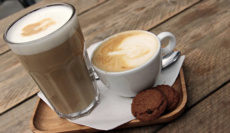 Two cups of coffee – one in a tall glass and the other in a white coffee cup – sit on a small wooden tray, on a larger wooden table.