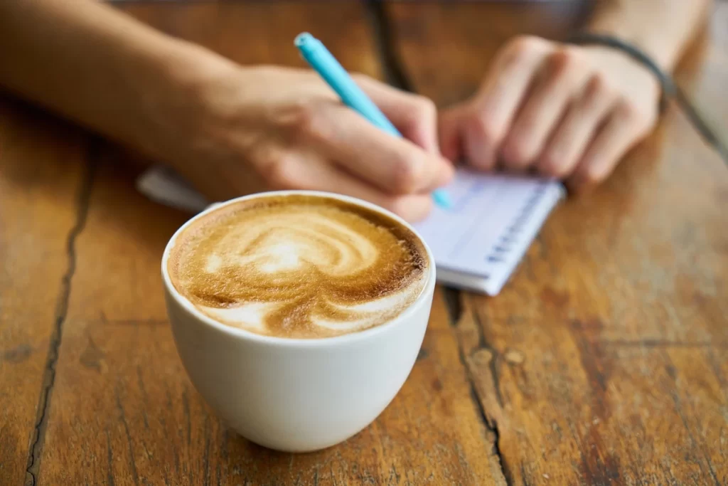 A full coffee cup (with swirly froth) sits on a wooden table. Behind the cup, a woman writes on a notepad with a pale blue pen.