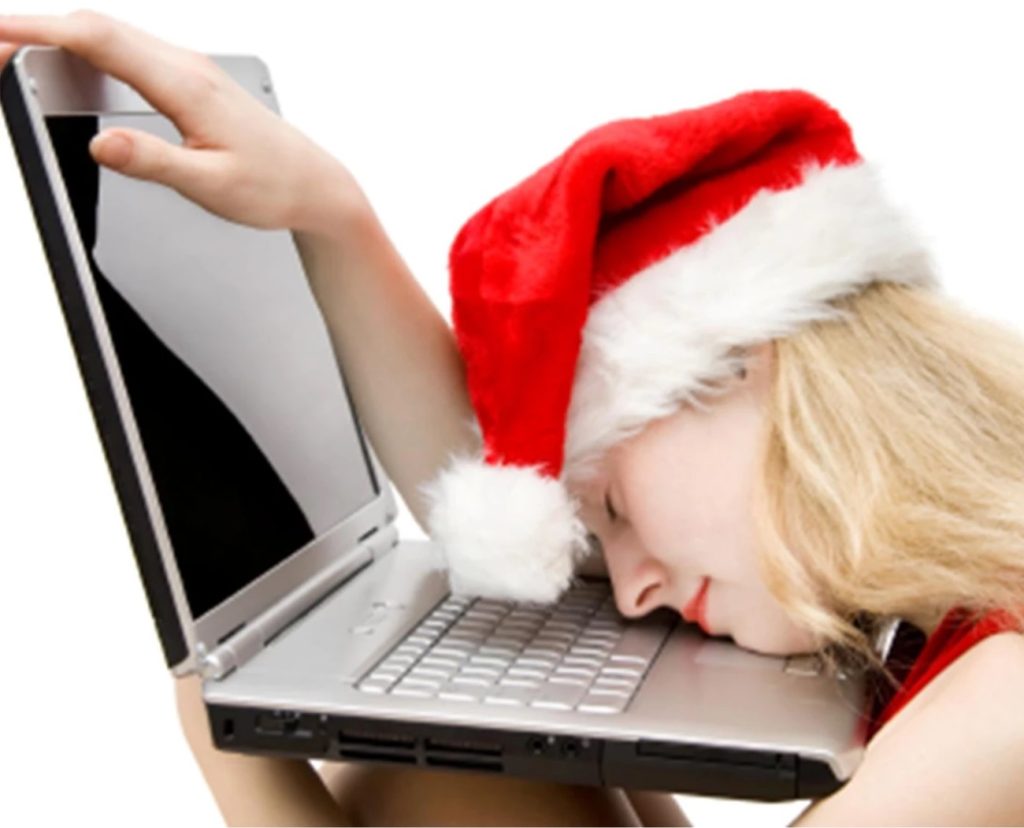 A blonde woman wearing a santa hat rests her head on the keyboard of an open laptop.