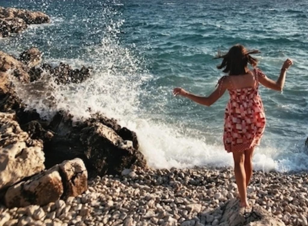 A woman in alight red and white sundress tiptoes across rocks towards crashing ocean waves.