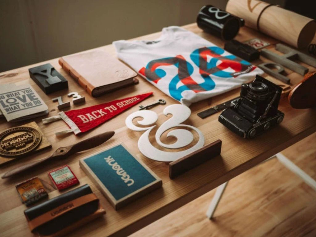 A close-up of a wooden table covered neatly by creative objects such as printing tools, old scissors, a camera, a notebook, etc.