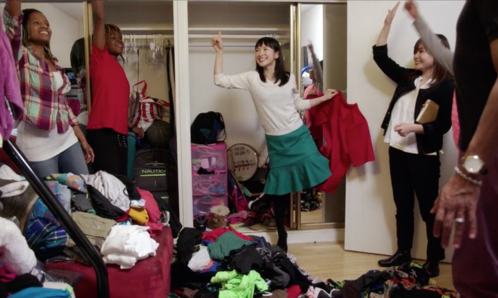 A scene from Marie Kondo's Netflic series, Sparking Joy. Marie is posed in front of an empry cupboard with it's contents on the floor. People stand either side watching Marie. She is smiling - an arm and a leg in the air.