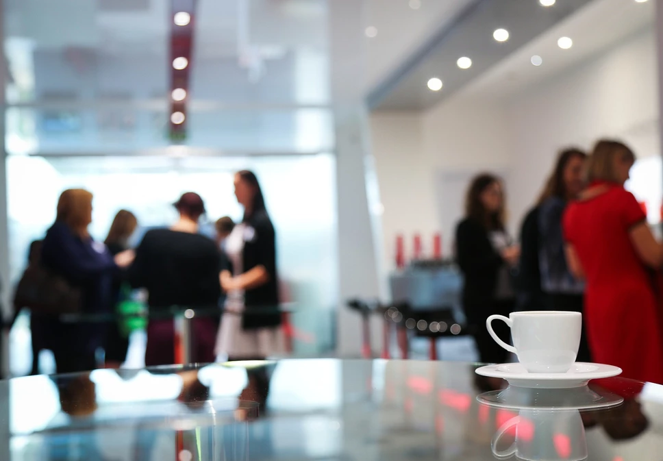 A group of women migle around a conference room. A white coffee cup sits on a table in the foreground. the picture is slightly out of focus.