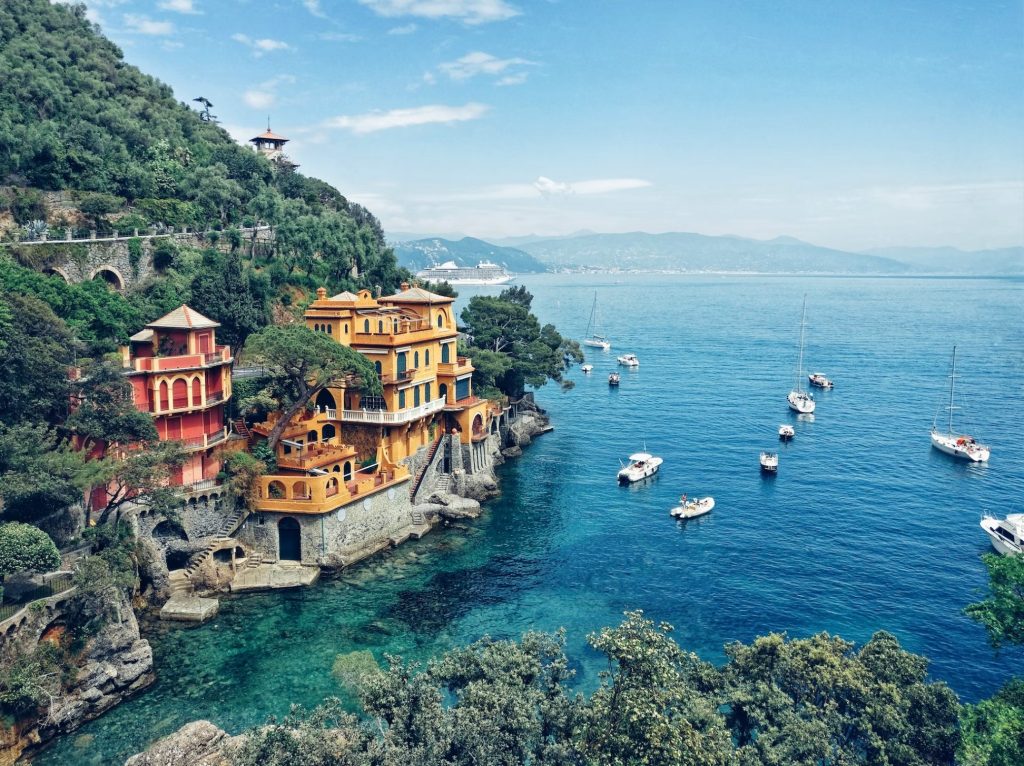 A wide view of part of the Portofino coast, with a few colour buildings set into a cliff on the left. The water is very blue. There are half a dozen or so boats. There are blue mountains on the horizon.