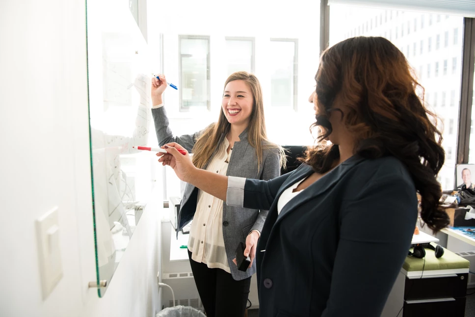 Two women holding markers stand and talk in front of a whiteboard.