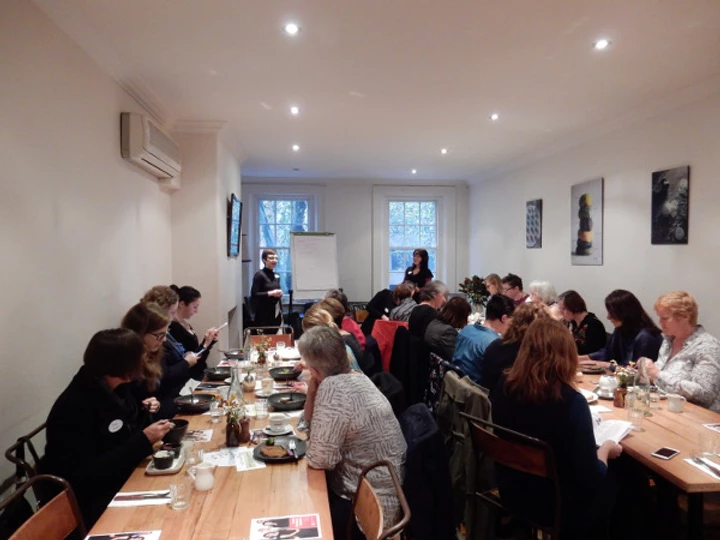 A room full of women attend a Take on Board breakfast. Thy sit at two long breakfast tables while listening to the guest presenters who are standing at the front of the room beside the windows.